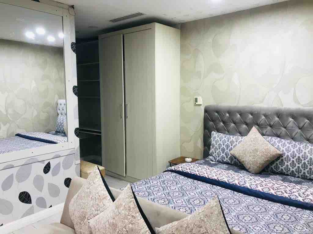 The studio features a spacious sleeping area with a tufted headboard and decorative pillows well-placed on the bed. A mirrored wardrobe reflects the light, and a subtle patterned wallpaper adds to the decor. Soft lighting enhances the cozy atmosphere.