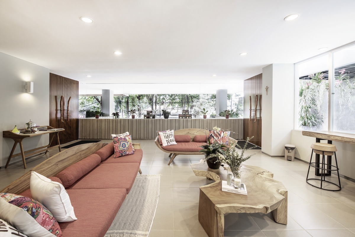 A spacious communal area is featured with light-toned seating arrangements, including two low couches adorned with decorative pillows. The room showcases large windows with views of greenery, and a natural wood coffee table is central to the space.