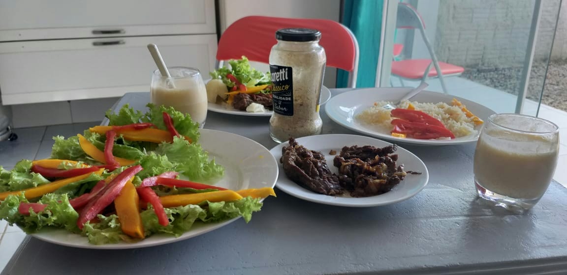 A table is set with a variety of dishes, including fresh salads garnished with colorful bell peppers. Bowls of rice and cooked meat are present, alongside beverages in clear glasses. The setting is well-lit, with a modern, minimalist aesthetic reflected in the background.