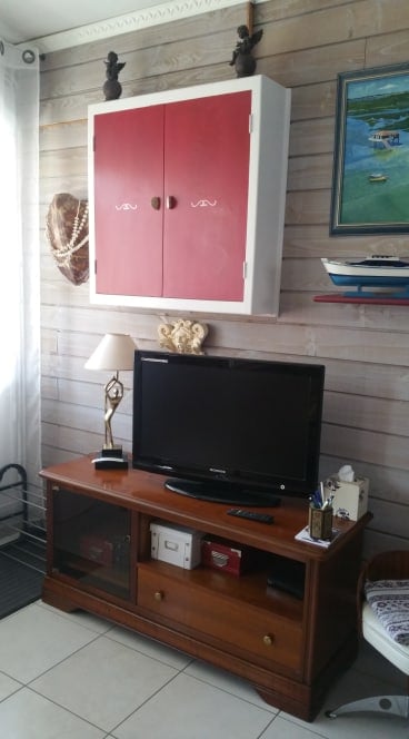 A wooden entertainment center displays a television beneath a wall-mounted cabinet with red doors. A small lamp and decorative items are positioned on the surface, and a painting depicting a water scene is hung above. Light streams in from a nearby window.