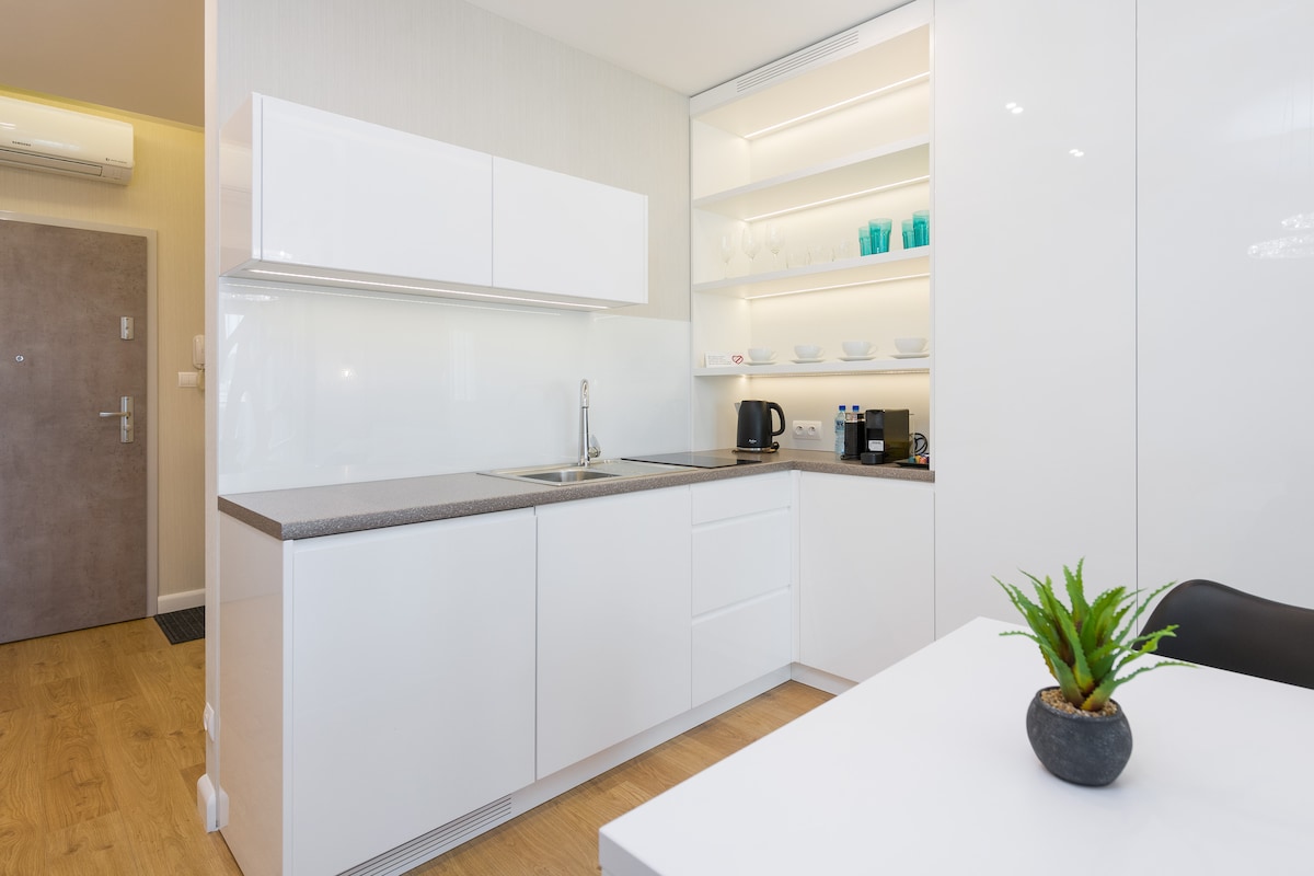 A modern kitchen space is showcased, featuring sleek white cabinetry and a light countertop. Clean lines and minimalistic shelves offer ample storage. Kitchen appliances include a kettle and coffee machine, while a small plant adds a touch of greenery to the environment.