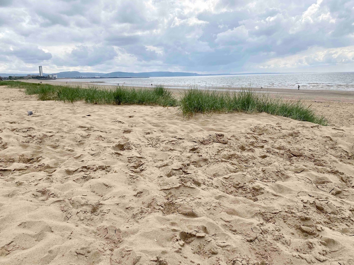A sandy beach is depicted, with gentle dunes covered in grass at the forefront. The tranquil sea stretches out to the horizon, under a sky adorned with soft clouds. The scene captures a serene coastal atmosphere, inviting exploration and relaxation.