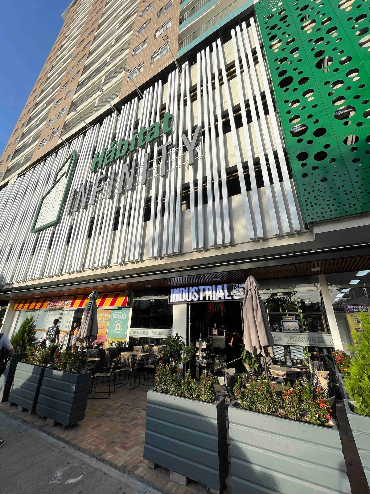 The exterior of a modern building is shown, featuring a large green sign that reads 'Hotel Infinity.' The structure has distinct architectural elements with metal slats and greenery integrated into the façade. Outdoor seating areas are visible, accompanied by potted plants.