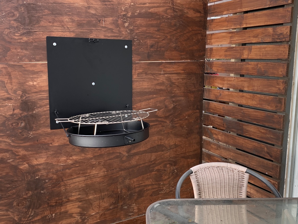 An outdoor grilling area features a wall-mounted grill, designed for easy access. A circular cooking grate is positioned above a black base, complemented by a wooden backdrop. A glass table and a chair with a woven seat are visible in the foreground.