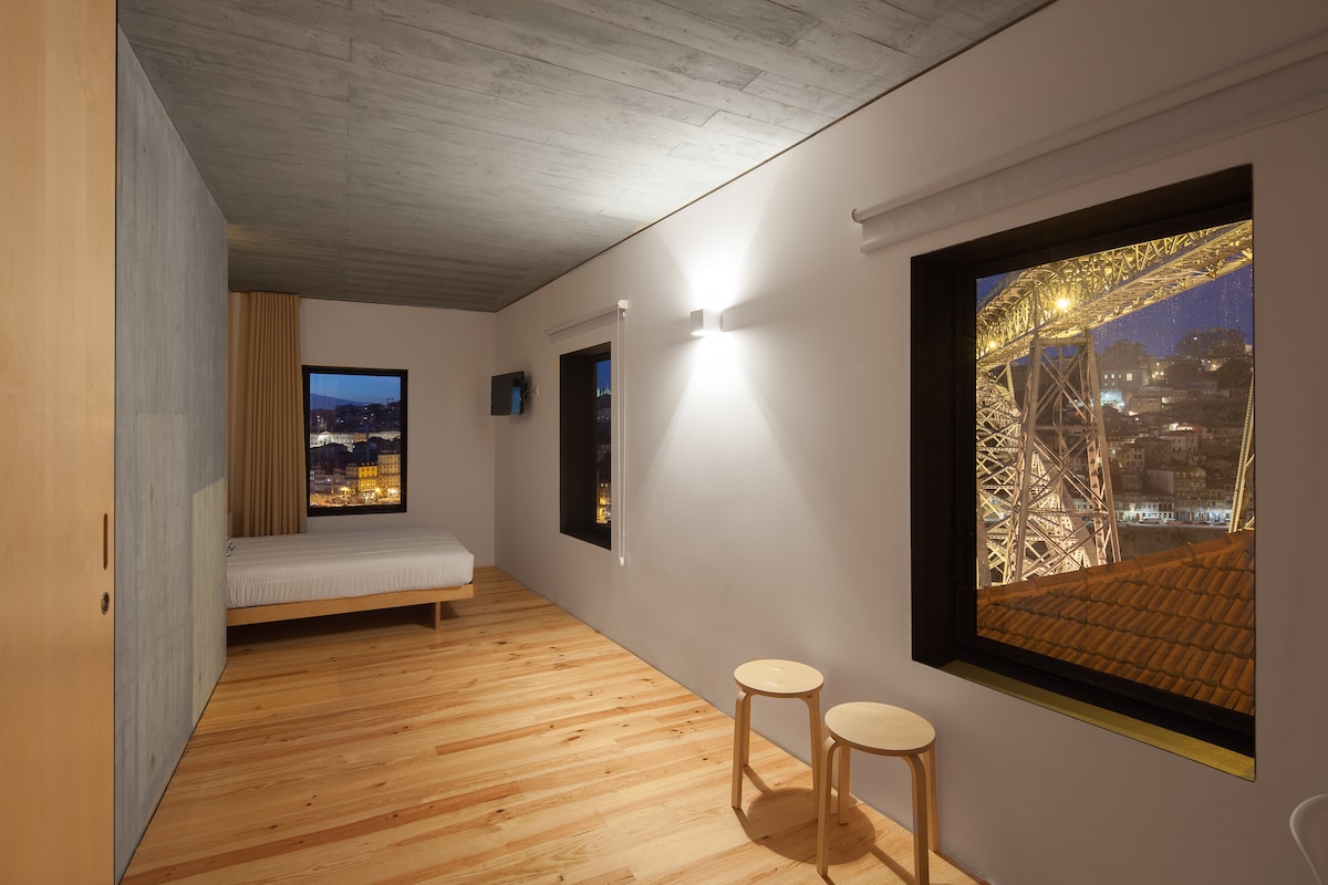 A spacious room is highlighted by warm wooden flooring and an expansive window offering views of the illuminated D. Luís bridge at night. A simple bed is positioned against the wall, complemented by two wooden stools in the foreground. Soft lighting enhances the modern design.