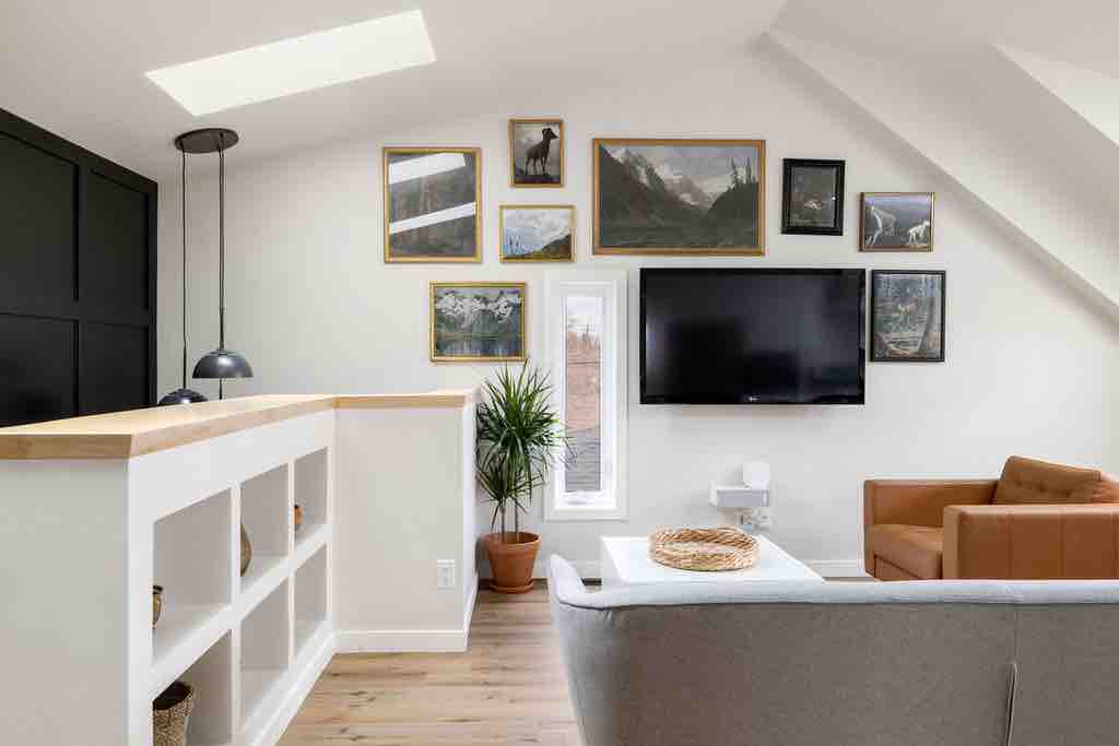 A modern living space is showcased with light-colored walls and a sloped ceiling. A large wall features a collection of framed nature artwork. A flat-screen TV is mounted above a light coffee table, accompanied by a gray sofa and a tan armchair, adding comfort to the area.