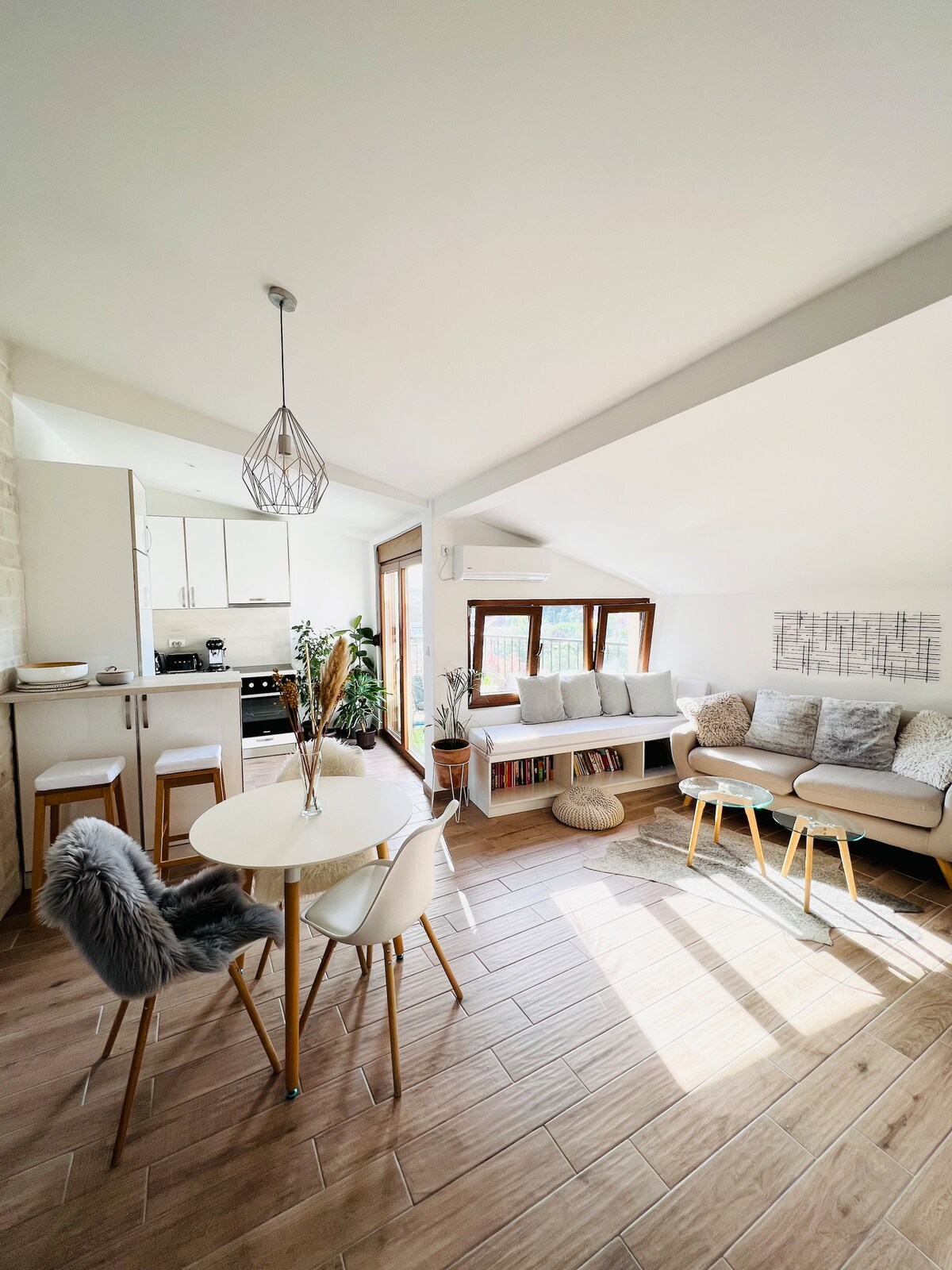 A well-lit living space is shown, featuring a cozy seating area with a light-colored sofa and decorative cushions. A dining table with four chairs is positioned nearby, complemented by soft decor elements. The kitchen area is visible in the background, with modern cabinetry and appliances.