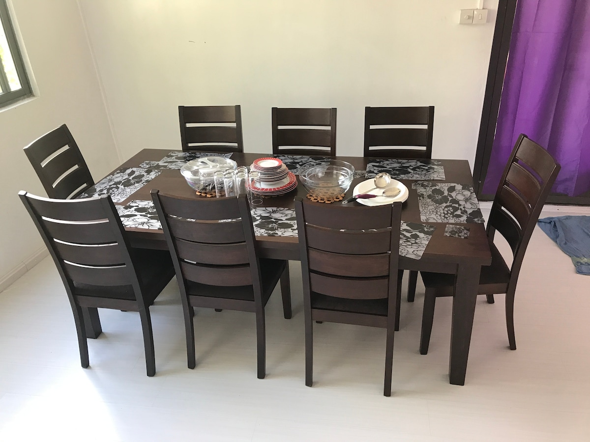 A modern dining table is presented with eight dark wood chairs arranged around it. The table is set with transparent glassware and a variety of plates, and is adorned with a decorative table runner featuring a floral pattern.