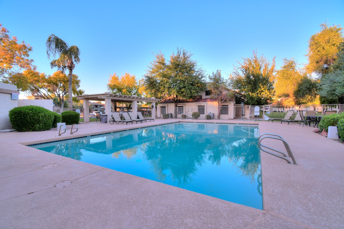 A sparkling outdoor pool is surrounded by lounge chairs and lush landscaping. Sunlight reflects off the water, and palm trees can be seen in the background. A shaded area with seating provides a place to relax near the poolside.