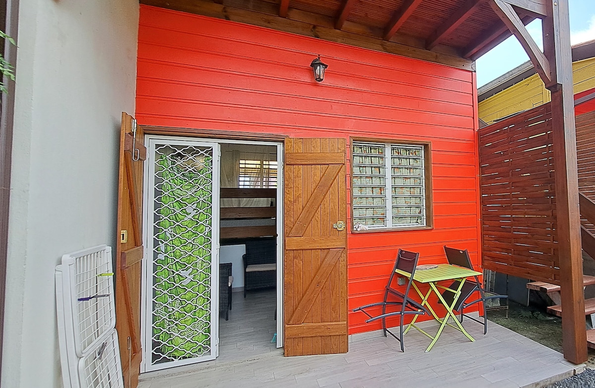 An exterior view of a vibrant red wooden structure showcases a welcoming entrance. Adjacent patio furniture includes a small green table and two chairs, inviting relaxation. Large windows allow natural light to filter into the interior, which is partially visible through an open door.