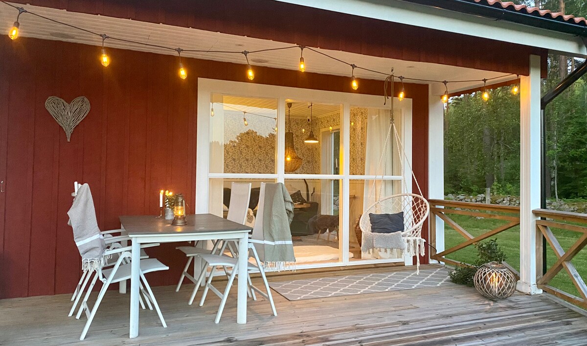 A spacious veranda is shown, featuring a wooden deck illuminated by warm string lights. A dining table with chairs and a cozy hanging chair create inviting seating areas. Large glass doors lead into the indoor living space, surrounded by natural views.
