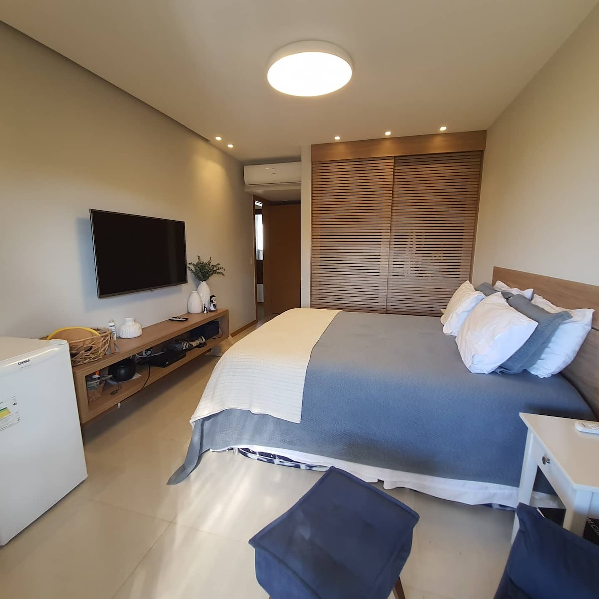 A modern bedroom is presented, featuring a spacious bed adorned with light blue and white linens. A wall-mounted television is positioned above a low media console, while a small white nightstand complements the decor. Natural light enters through a nearby window, enhancing the room's ambiance.