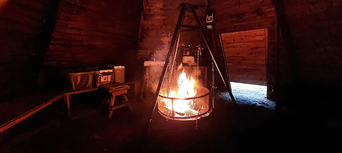 A cozy indoor space with a traditional fire pit at the center, surrounded by wooden walls. The flame flickers brightly, illuminating the area, while a wooden table and benches are positioned nearby. An entrance is visible, providing a glimpse of the natural surroundings outside.