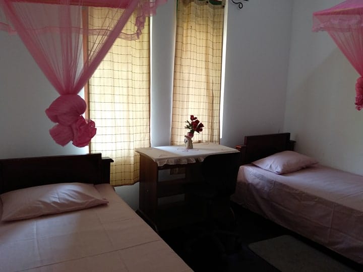 A room featuring two single beds arranged beside each other, each adorned with light pink bedding. A small desk sits between the beds, holding a vase with flowers. Soft light filters through the sheer curtains, enhancing the calm atmosphere.