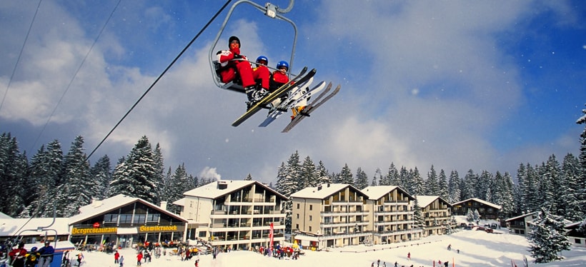 A ski lift is in operation, transporting individuals dressed for winter sports. Below, snow-covered terrain features several chalet-style buildings, surrounded by tall evergreen trees. The scene showcases the winter landscape with a clear blue sky and gentle clouds.