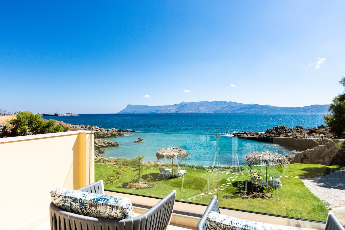 A view of the tranquil sea and distant mountains is offered from the villa's veranda. Two outdoor sitting areas, protected by umbrellas, are set on lush green grass, leading to a private beach cove. The clear blue sky enhances the serene atmosphere.