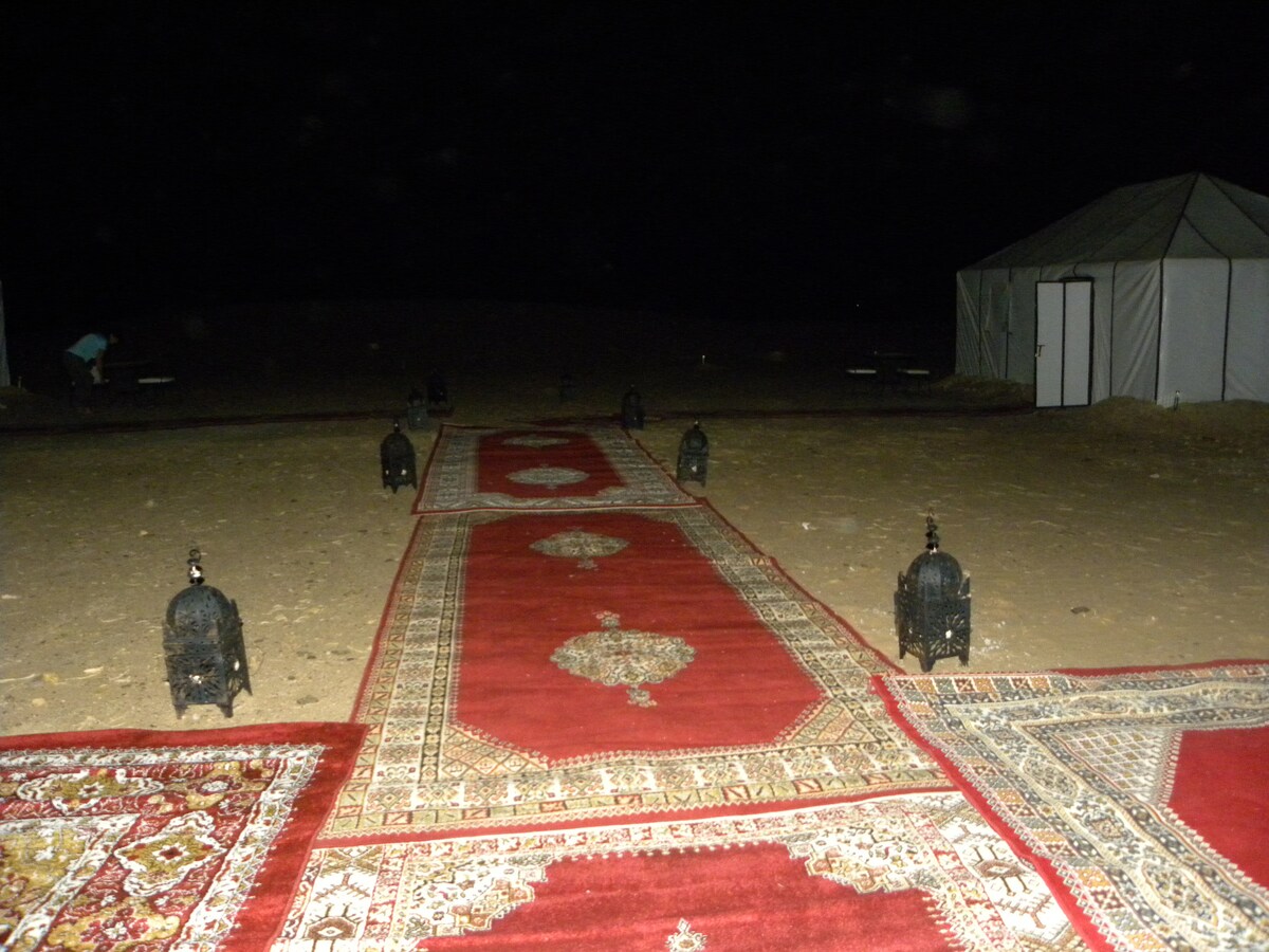 A pathway of ornate rugs is visible, leading toward tents in the desert at night. The rugs are bordered by low, decorative lanterns, providing ambient lighting. The surrounding area is characterized by sand and the silhouettes of distant dunes under a starlit sky.