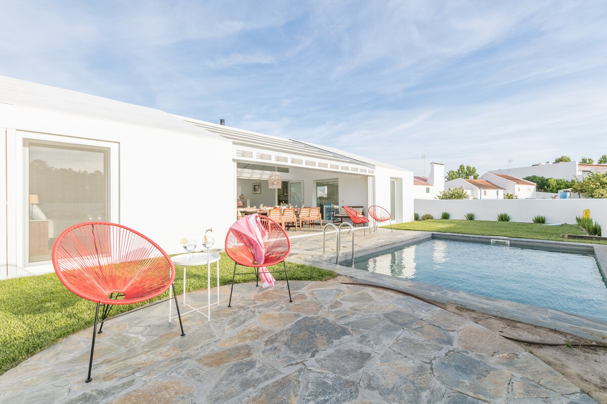 A spacious outdoor area features a clear swimming pool bordered by a stone patio. Two bright red chairs are positioned beside a small table, inviting relaxation. The white exterior of the house complements the lush green lawn and is enhanced by large sliding glass doors.