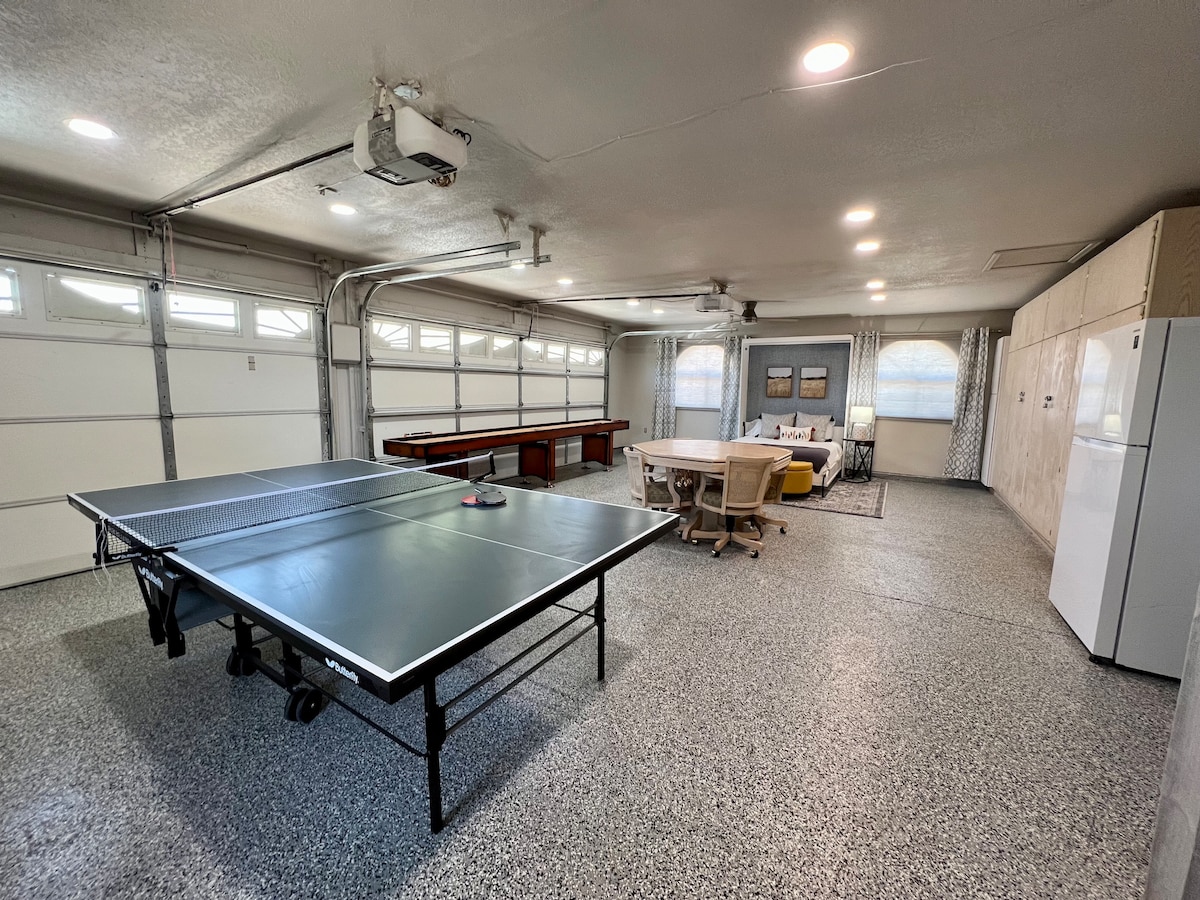 A spacious garage area is shown, featuring a large ping pong table positioned centrally. A refrigerator stands in one corner, while a cozy seating area with a table and chairs can be seen in another corner, illuminated by overhead lights.