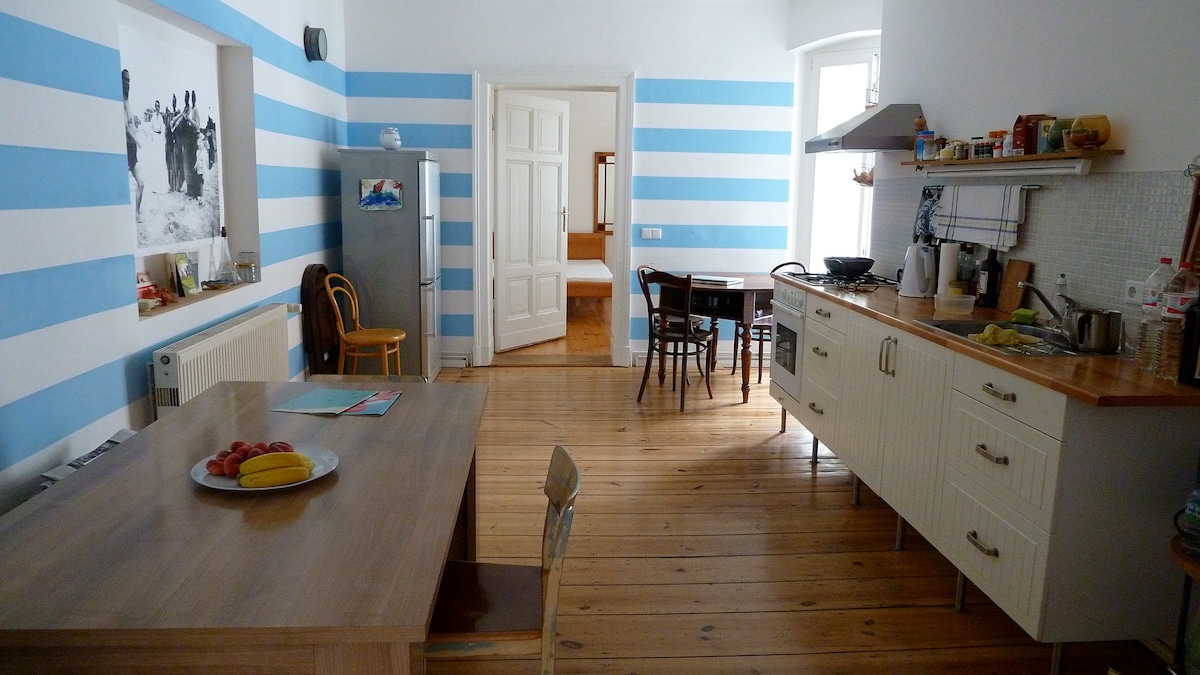 A spacious kitchen area features striped blue walls and wooden flooring. A dining table with chairs is positioned alongside a modern countertop equipped with cooking essentials. A refrigerator and various kitchen items are visible, while a doorway leads to another room in the background.
