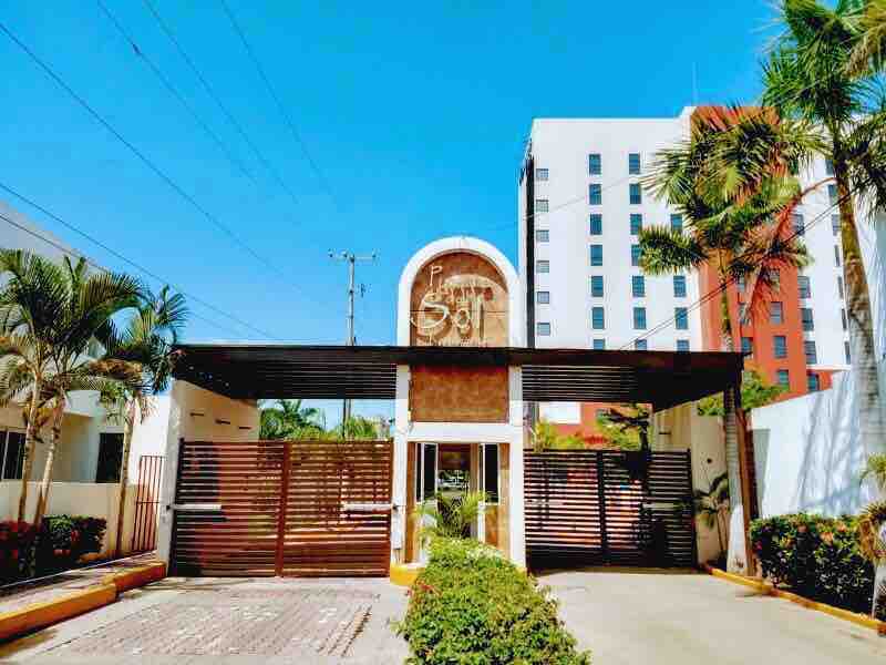 The entrance of a gated community features a welcoming arch with the name 'Casa del Sol' prominently displayed. Lush greenery and decorative plants frame the entrance, while a clear blue sky sets a bright backdrop for the modern architecture.