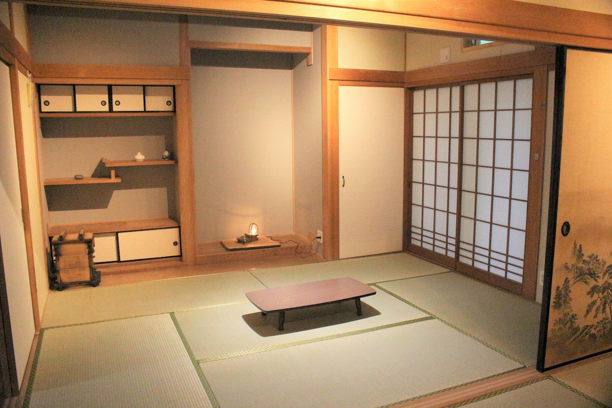 A traditional Japanese tatami room features a low table at its center, surrounded by light-colored tatami mats. Shoji doors offer a view of a softly lit area, while a simple shelf displays decorative items, contributing to a calming and minimalistic design.