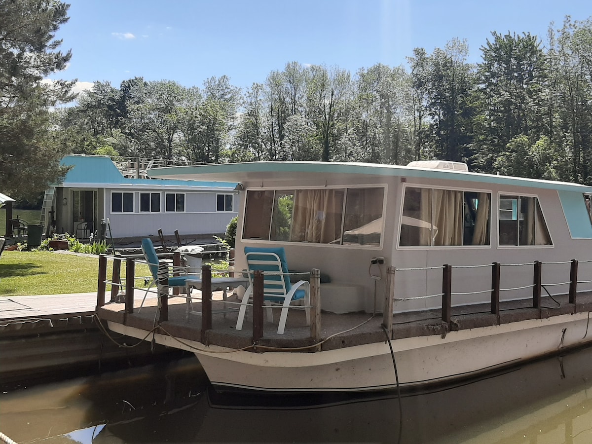 A floating cottage is docked at the canal, surrounded by lush greenery. Comfortable seating is visible on the front deck, while the main structure displays large windows offering natural light. The serene waters reflect the charming setting and adjacent building.