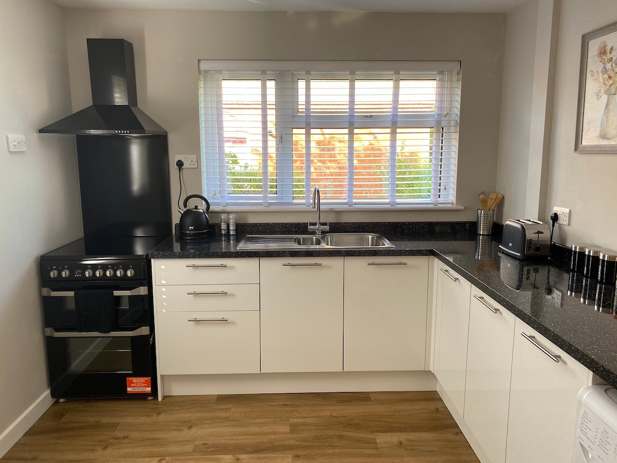 The kitchen features modern white cabinetry paired with a sleek black countertop. An oven and stovetop are situated on one side, while a stainless steel kettle and toaster are placed on the countertop. A window with white blinds allows natural light to fill the space.