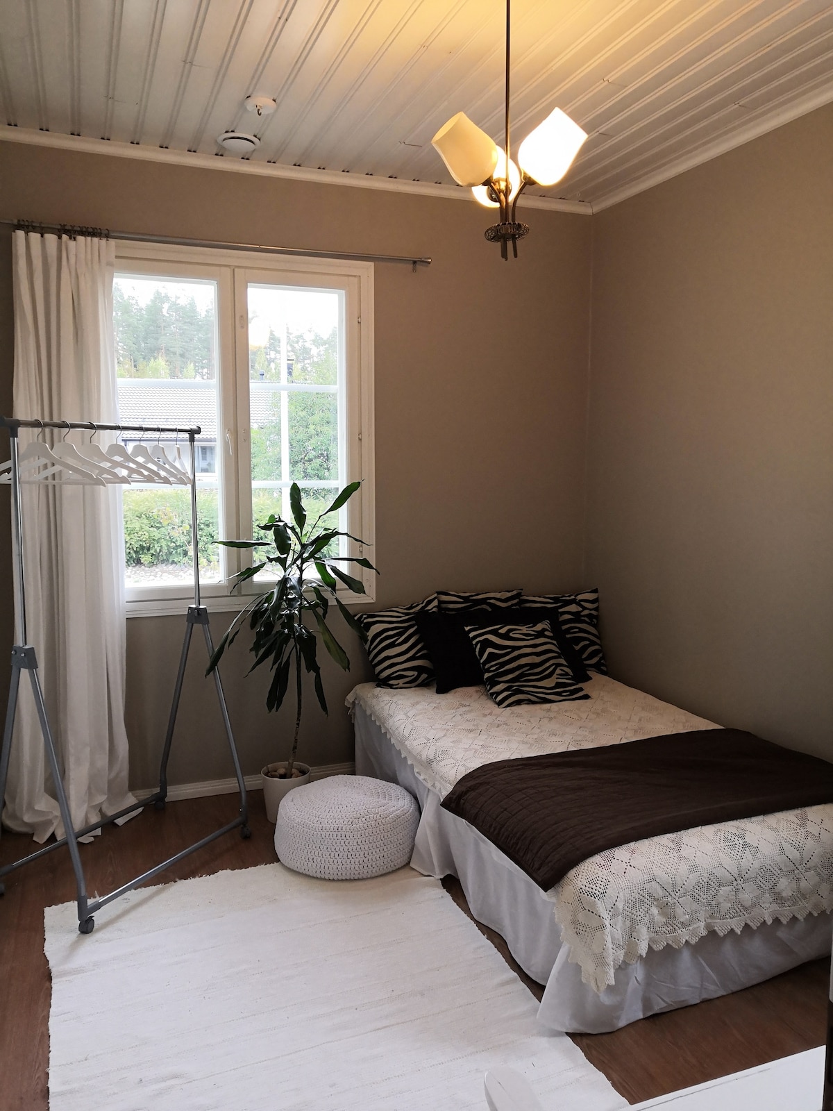 A cozy room features a single bed dressed in neutral linens with decorative cushions. A standing clothes rack and a small, round pouf are included. Natural light fills the space through a large window, complemented by sheer curtains and a leafy plant nearby.