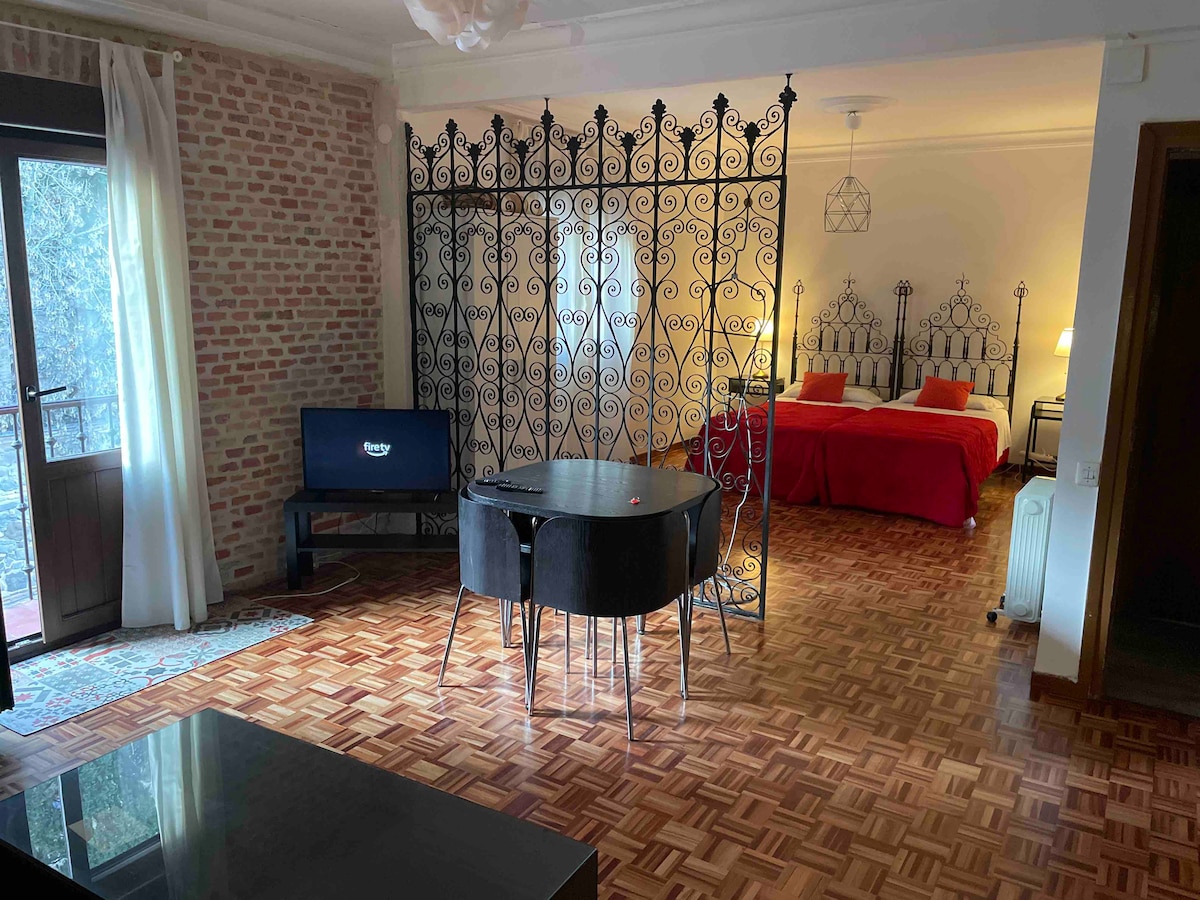 An open living space is divided by an ornate iron screen, revealing two spacious beds beyond. A round black dining table sits in the foreground, accompanied by sleek chairs. Natural light filters through the glass door, highlighting the warm wood flooring and exposed brick wall.