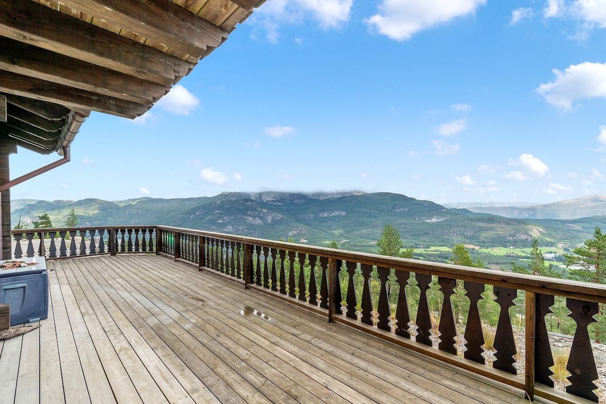 A spacious wooden deck offers panoramic views of the surrounding mountains and valleys. The deck features a balustrade with intricate detailing, enhancing the outdoor space. A hot tub is positioned on one side, inviting relaxation while enjoying the scenic landscape.