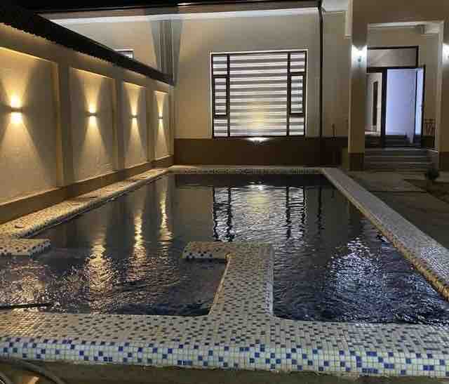 A tranquil swimming pool with a mosaic-tiled border is featured in a softly lit area. The water reflects the ambient light from the surrounding walls, and large windows allow for natural illumination, enhancing the serene atmosphere.