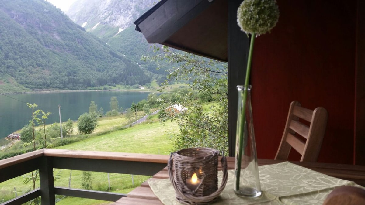 A balcony table is set with a single flower in a slender vase and a woven basket containing a candle. The view reveals green hills descending to a tranquil fjord, framed by towering mountains under a cloudy sky.