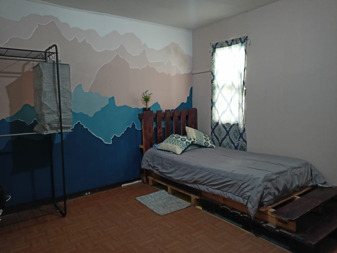 A room features a low platform bed with a gray coversheet, complemented by decorative pillows. A folding clothing rack stands nearby, alongside a small area rug. A window with a patterned curtain allows natural light to illuminate the wall, which is adorned with a mountain-themed mural.