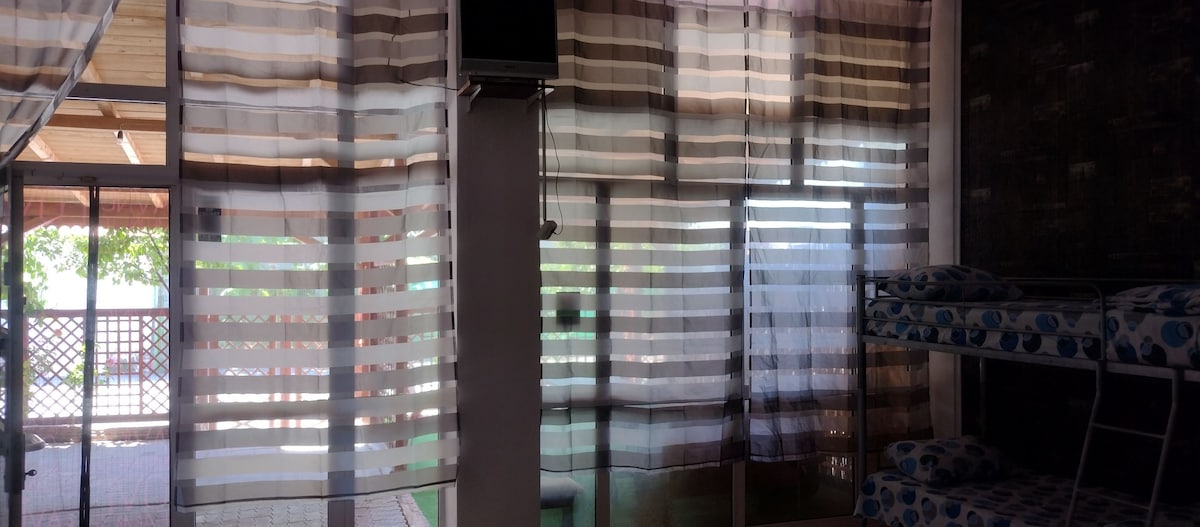 A bright room features large, sheer striped curtains that allow natural light to filter through. Opposite a bunk bed, a wall-mounted television is positioned above a white column. Sliding glass doors lead to an outdoor area, enhancing the spacious feel of the interior.