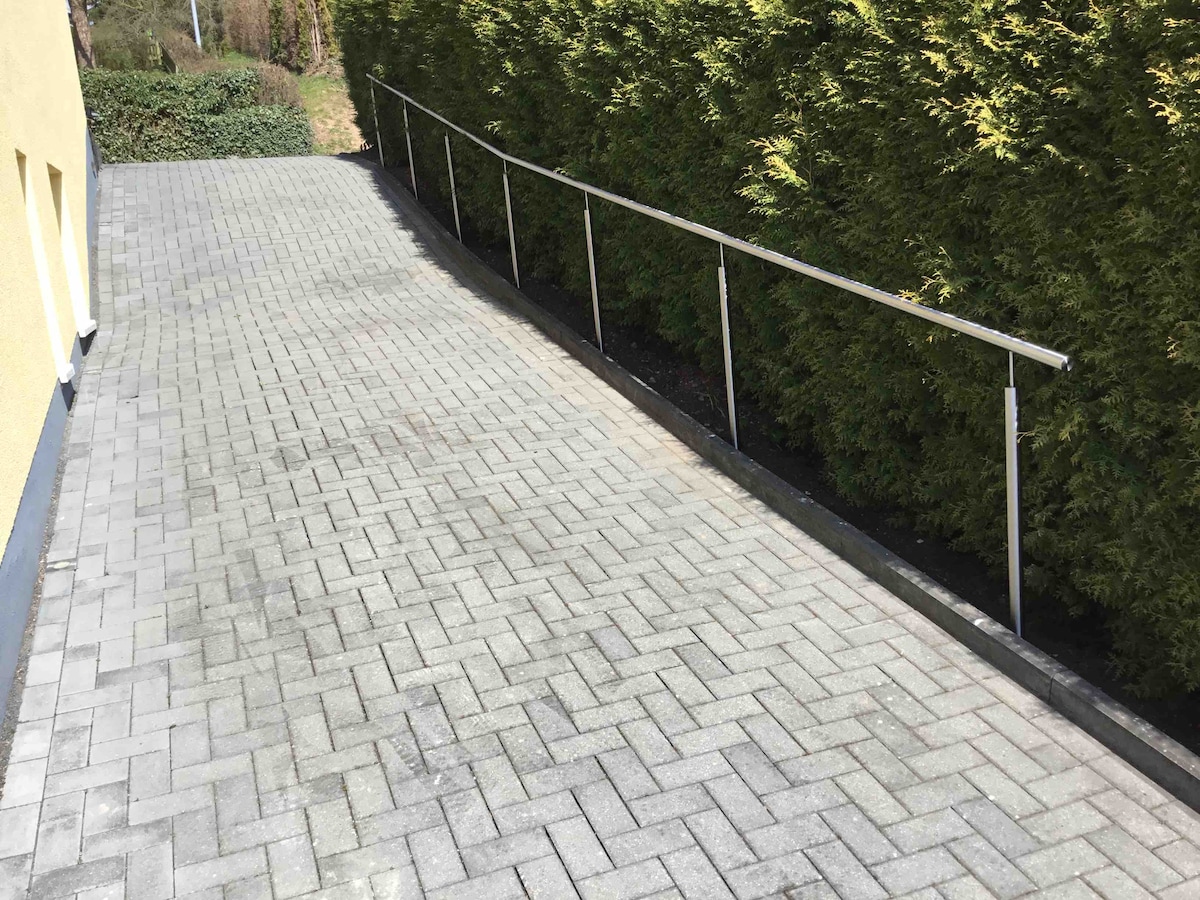 A clearly paved pathway leads gently downward, bordered by a vibrant green hedge on one side. A stainless steel railing offers support along the pathway. The ground is made of uniform bricks, providing a stable surface for navigation.