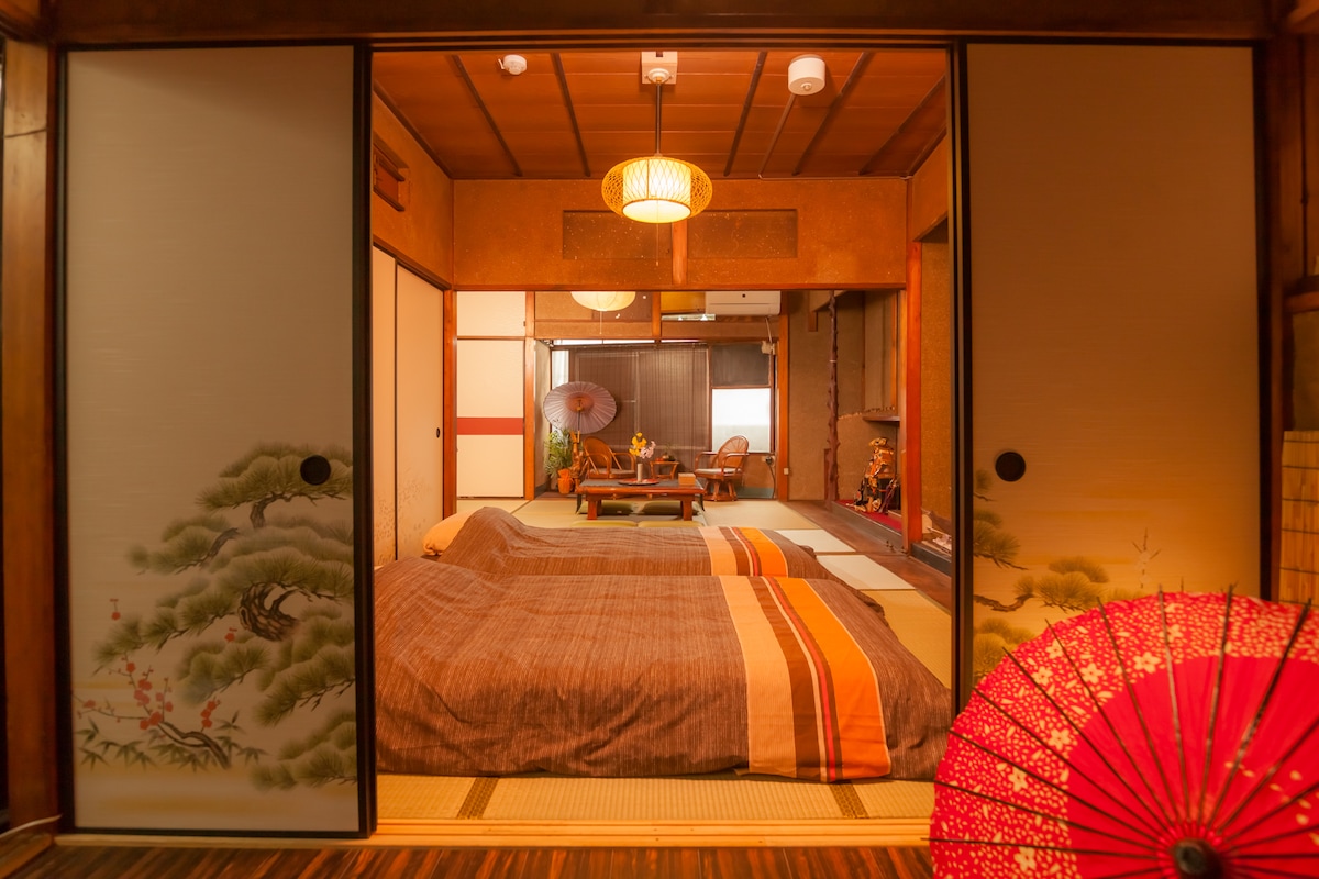Two futons are elegantly arranged on the tatami mat floor within a traditional Japanese room. Sliding paper doors, adorned with delicate artwork, separate the space. A warm light fixture hangs above, and a decorative red umbrella enhances the ambiance. A low table and seating area are visible in the background.