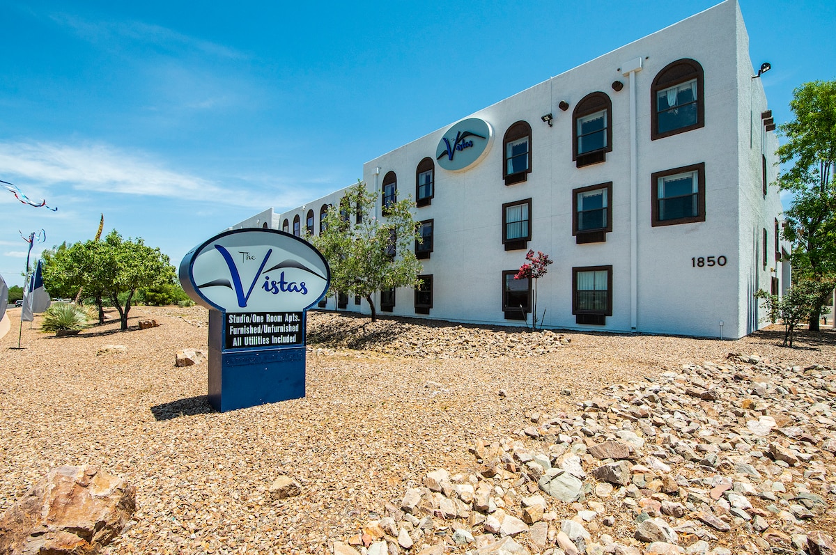 The exterior of The Vistas features a modern multi-story building with large windows and a prominent sign at the entrance. Surrounding shrubs and rocks create a natural landscape, contributing to the unit's welcoming presence in the area.