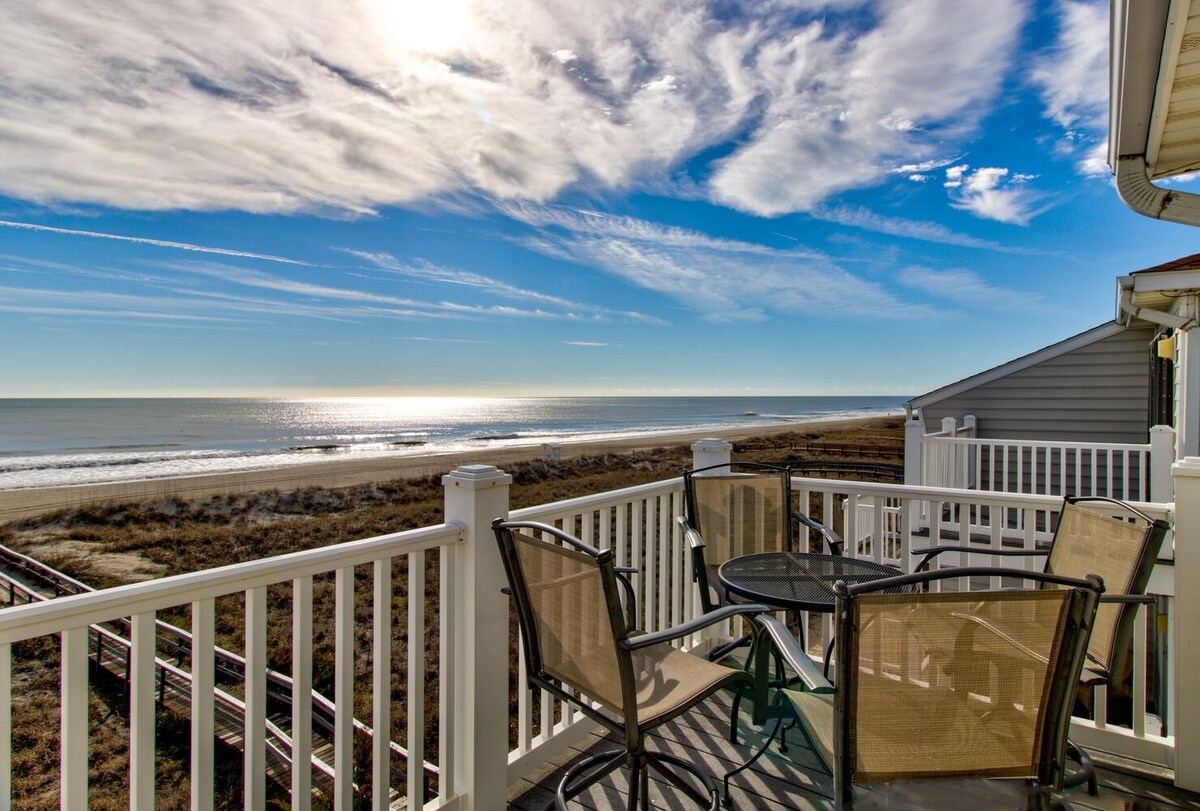 A spacious outdoor deck features a table and four chairs, overlooking a wide expanse of sandy beach and the Atlantic Ocean. The tranquil horizon is adorned with soft clouds and beams of sunlight, creating a serene atmosphere.