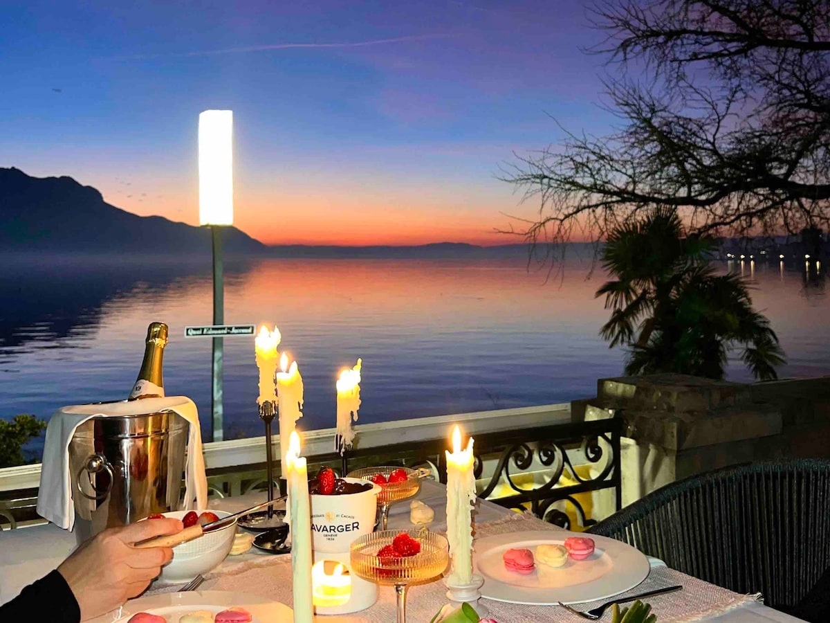 An outdoor dining setup is presented on a terrace, with a view of Lake Geneva at sunset. A table is elegantly arranged with candles, dessert items, and a champagne bottle. The horizon displays soft hues of orange and pink, reflecting on the calm water surface.