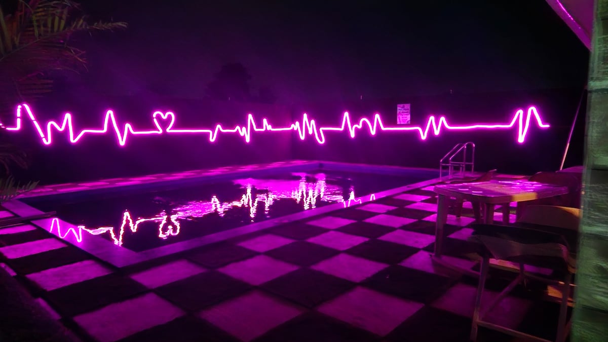 A swimming pool is illuminated by pink neon lights, creating a vibrant heartbeat design along the back wall. A checkerboard-patterned patio area complements the pool, while wooden tables and chairs are arranged nearby for seating. The nighttime setting enhances the ambiance.
