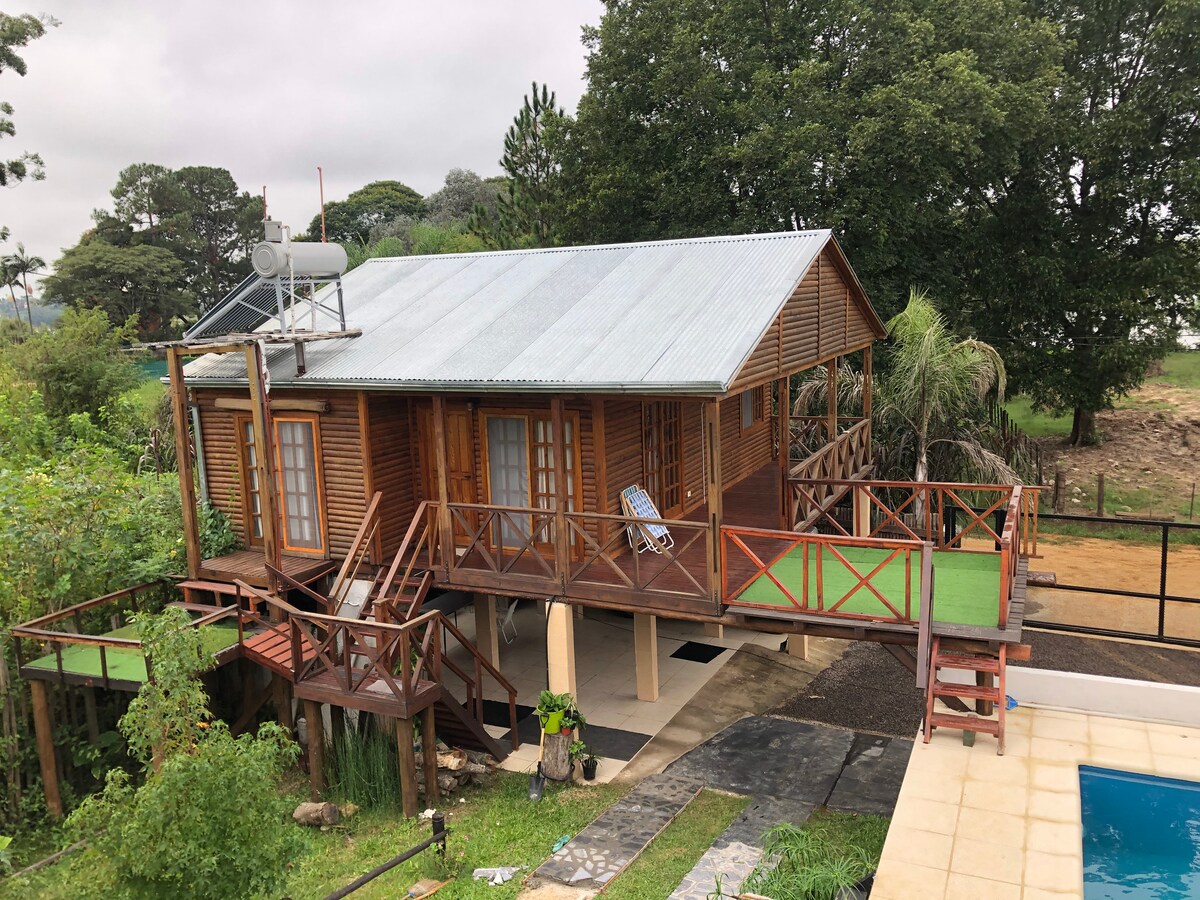 A wooden cabin is set on stilts within a lush green landscape. Expansive decks surround the cabin, featuring outdoor seating arrangements. A ladder leads up to the entrance, while a small pool is visible to the side. Trees and foliage enhance the serene environment.