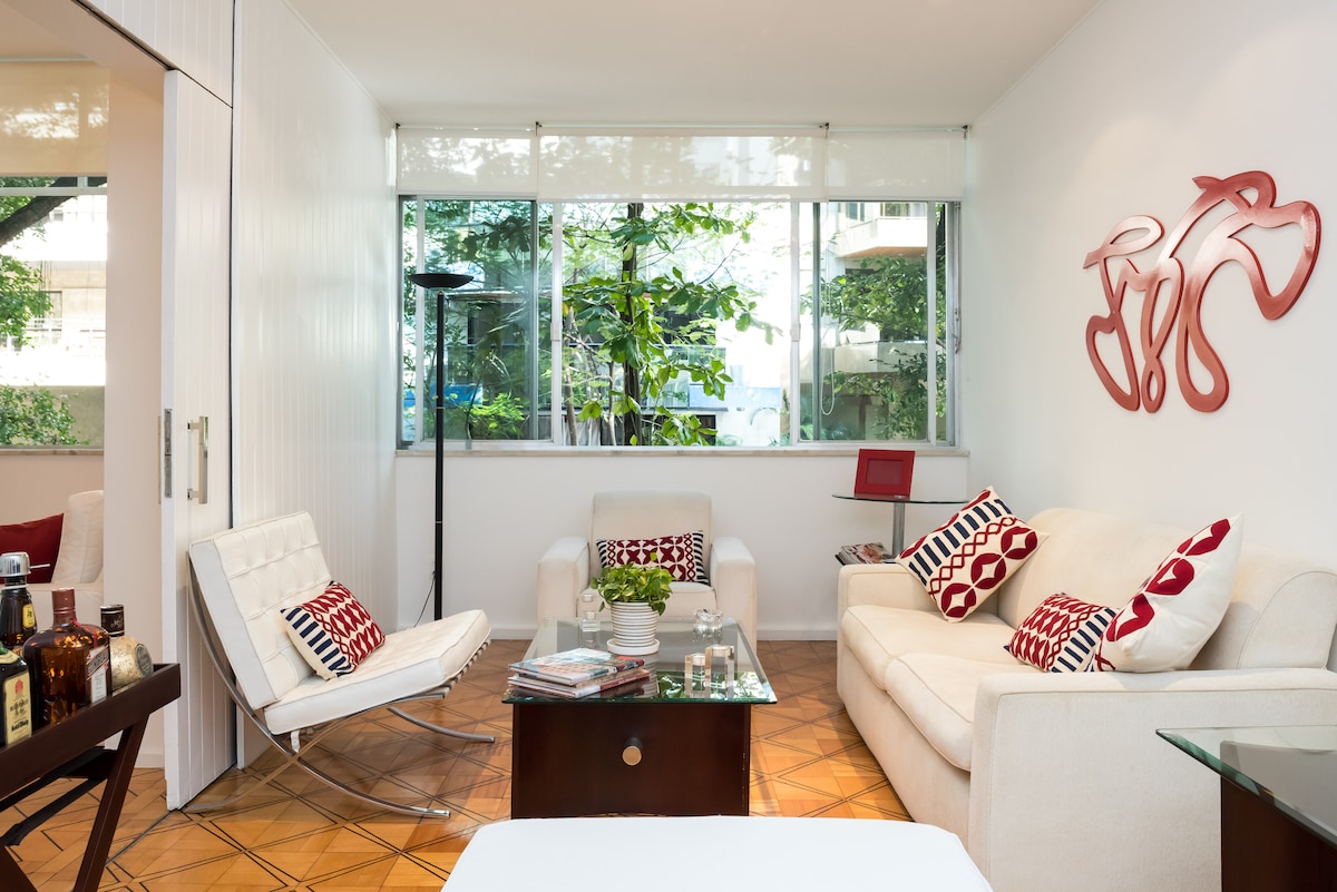 A bright living area features large windows allowing natural light to flow in, enhancing the greenery visible outside. Comfortable seating includes a white sofa and a modern chair. Decorative cushions with red patterns adorn the furniture, while a coffee table is centrally placed with simple decor.