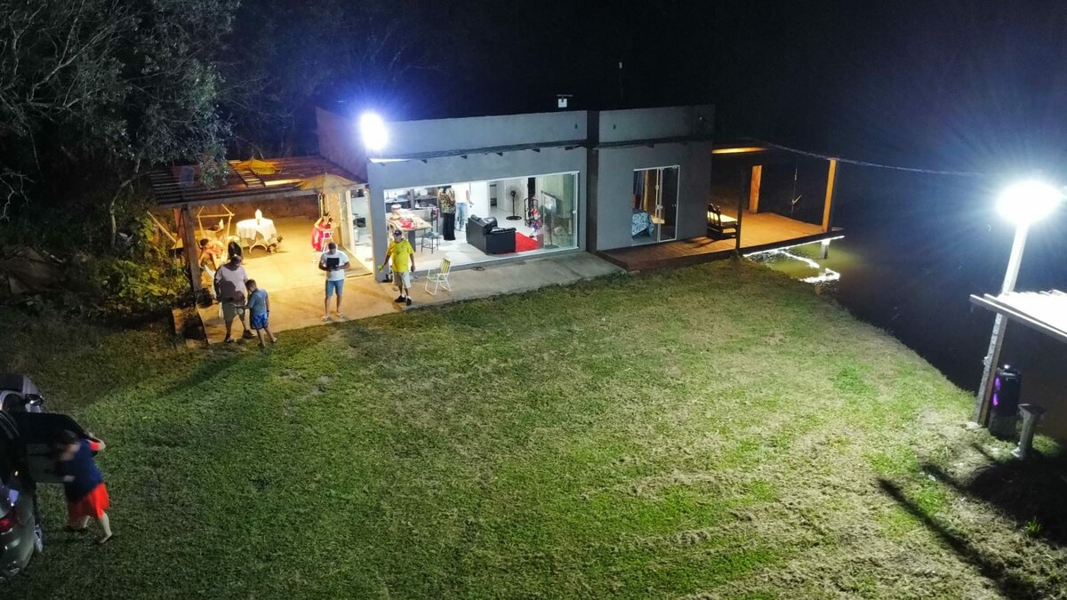 The image captures a spacious property at night, featuring a modern building with illuminated outdoor areas. Guests are seen enjoying the outdoors near a well-maintained lawn. A deck extends over the water, providing access to fishing, while the setting is enhanced by soft lighting from overhead fixtures.
