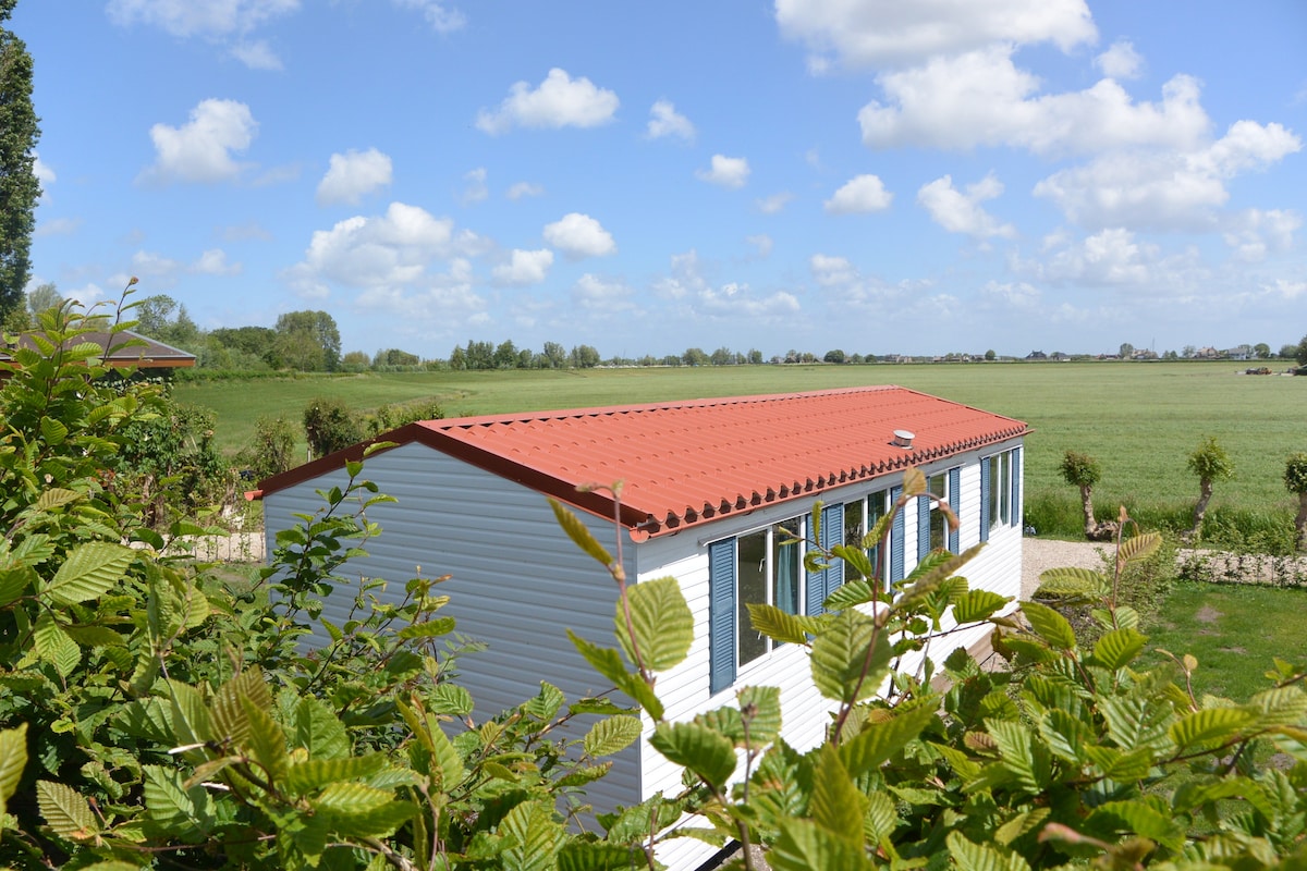 A modern vacation home is surrounded by lush greenery, featuring a red roof and multiple windows that invite natural light. Expansive views of open fields and the clear blue sky create a serene backdrop, emphasizing the property's location within a natural area.