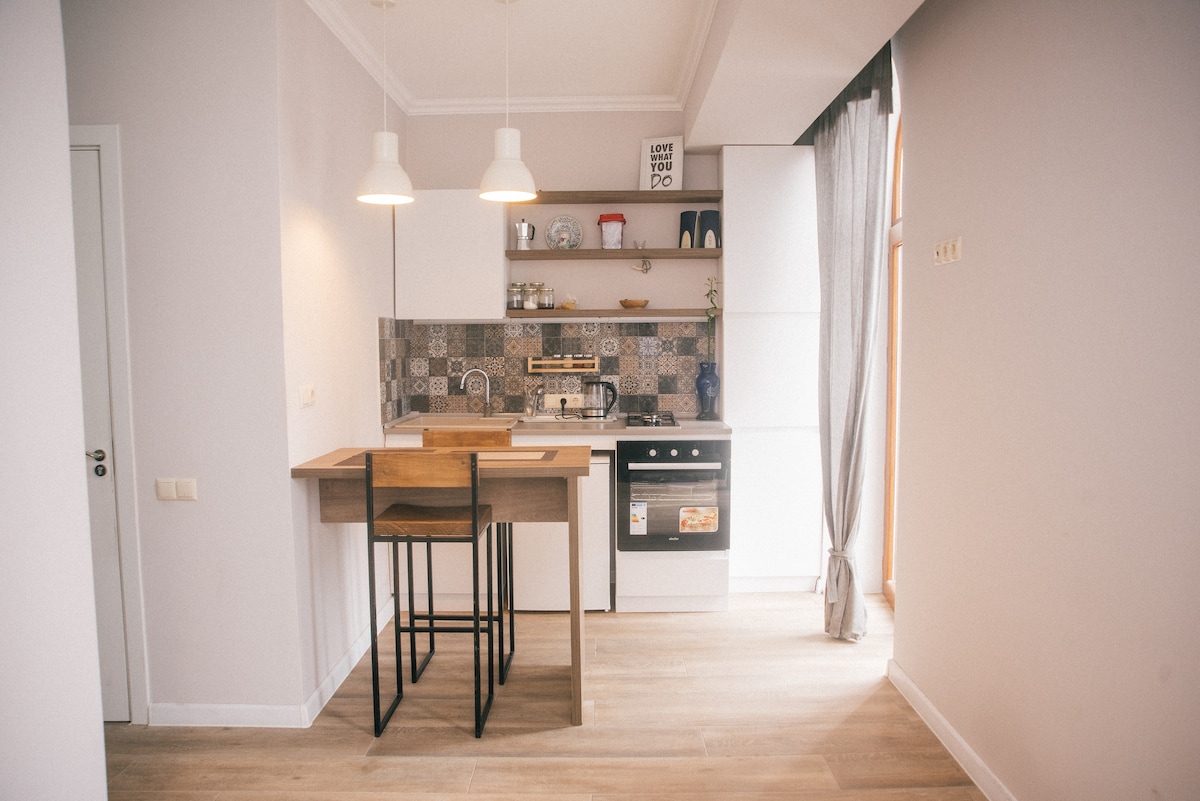 A modern kitchenette is highlighted with a tiled backsplash and equipped with a gas stove and cabinets. Two minimalistic bar stools are placed at a compact wooden table, enhancing the functional design. Natural light flows in through a nearby window adorned with sheer curtains.