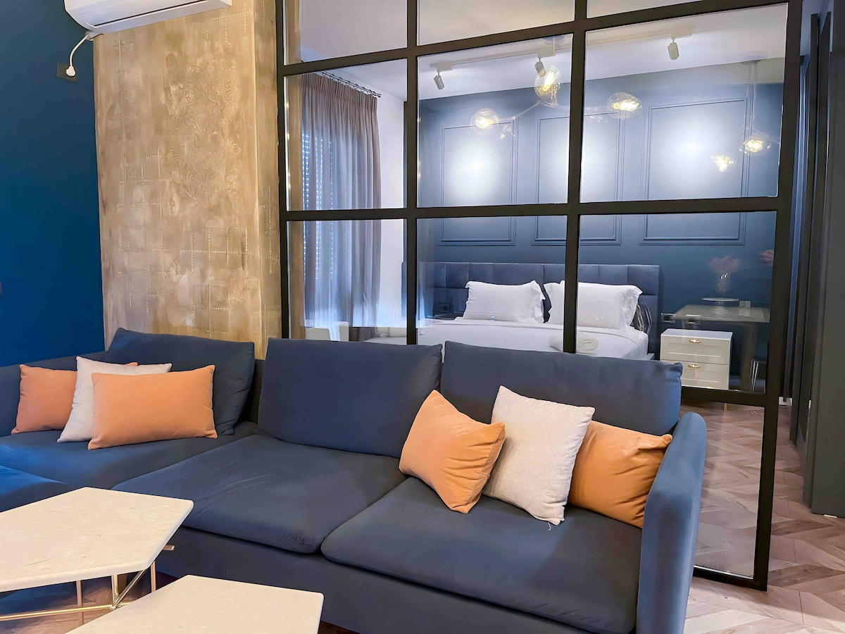 A stylish living area features a modern L-shaped sofa with orange and white accent pillows. Elegant glass partitions provide a view of a cozy sleeping area, where a spacious bed is complemented by soft lighting and contemporary decor.
