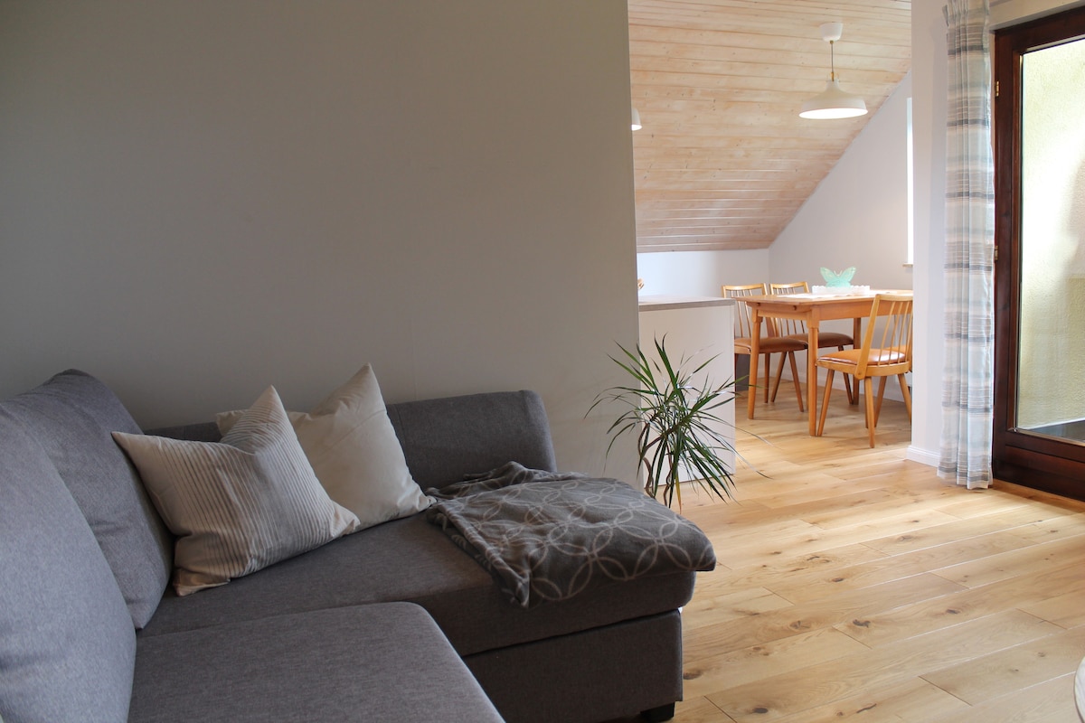 A sectional sofa features neutral-toned cushions and a decorative throw, positioned next to a light wooden floor. In the background, a dining area with a table for four is visible, with warm wooden chairs and natural light filtering through a window.