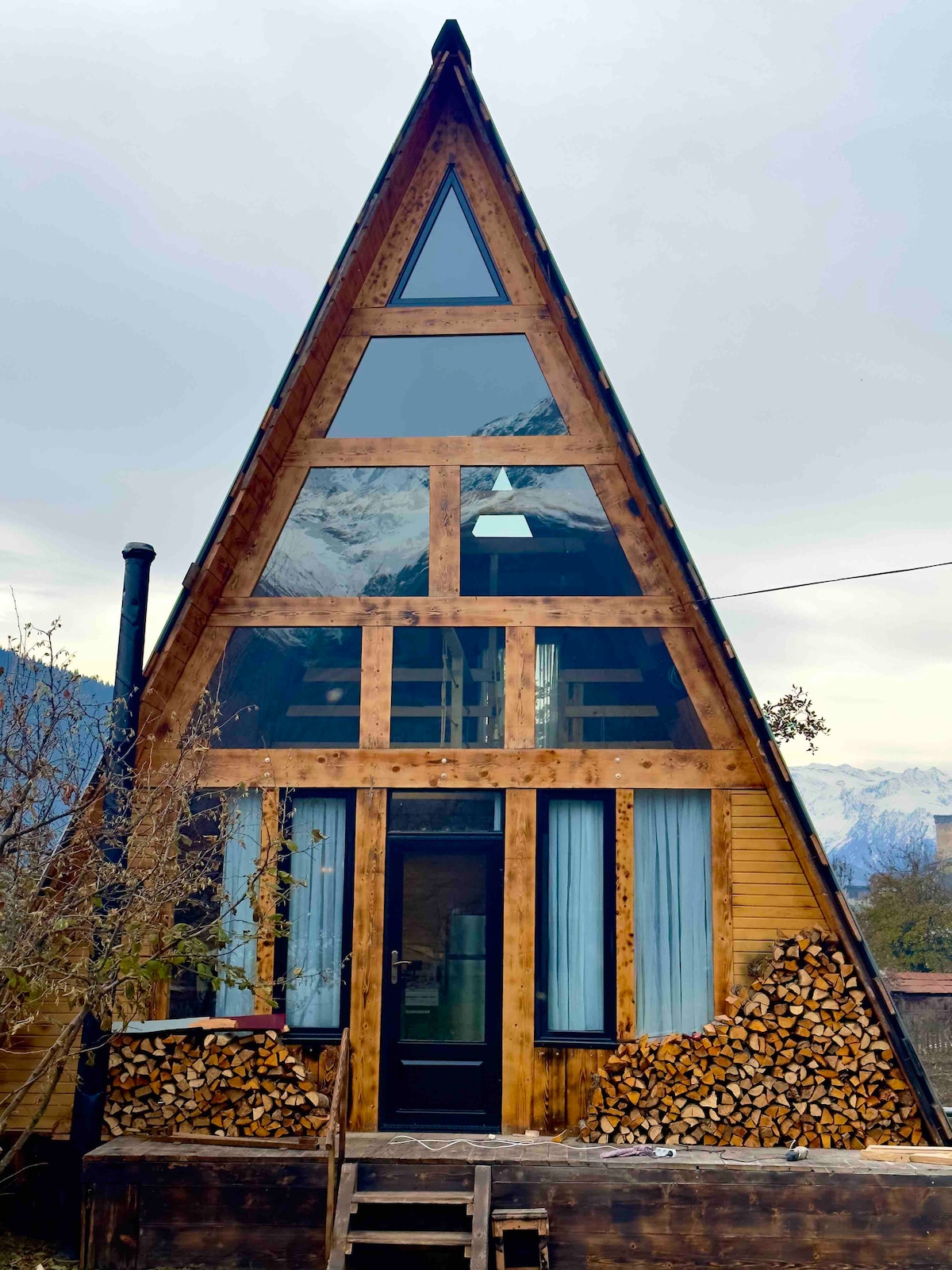 A triangular chalet features a wooden exterior with large glass windows that allow natural light to fill the interior. A neatly stacked pile of firewood is visible against the base, and steps lead up to the dark front door framed by light blue curtains.