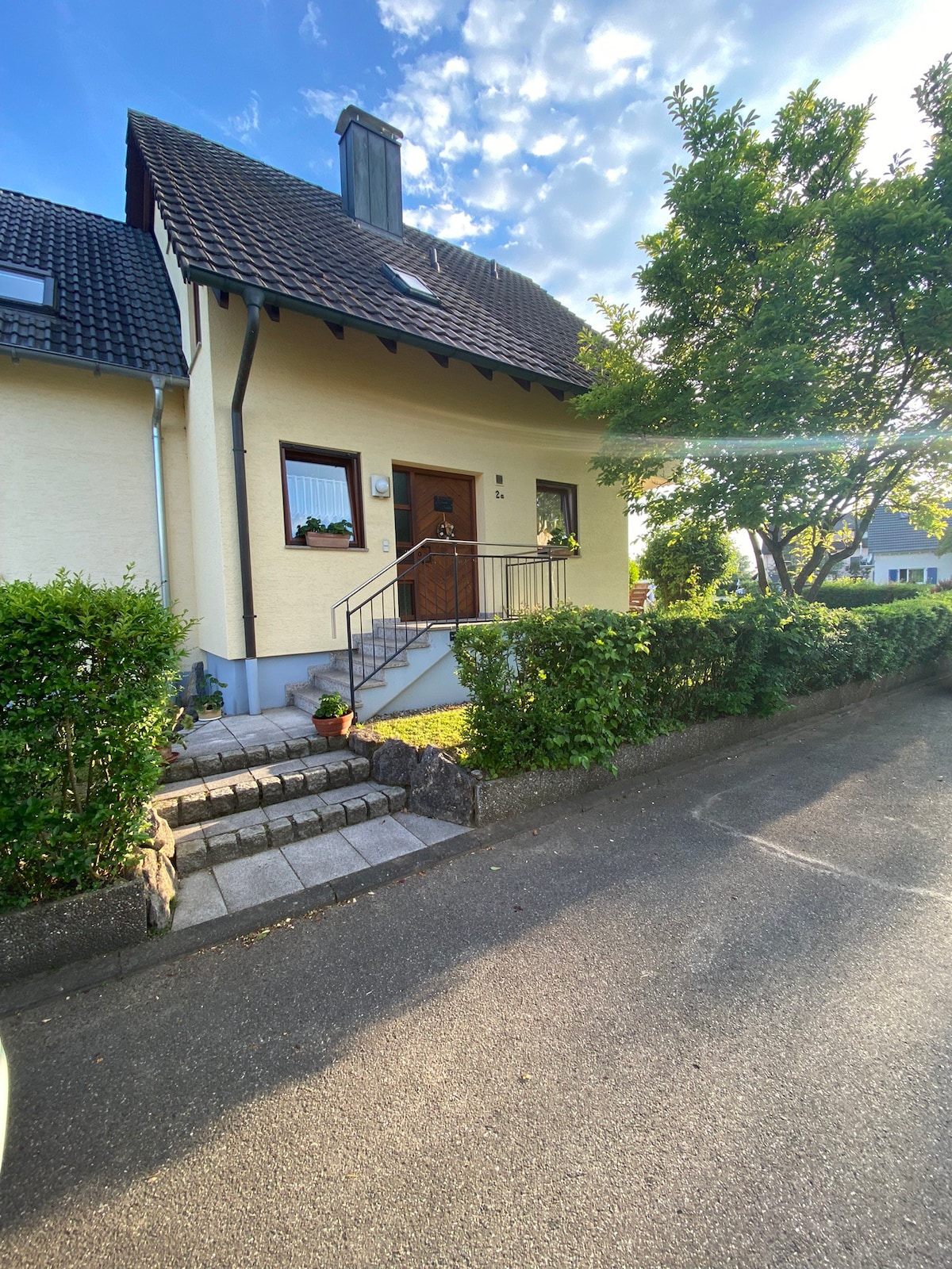 The front of the house is shown, featuring a welcoming center door with a small porch. A neatly trimmed hedge surrounds the entryway, with stone steps leading up to the door. Sunlight filters through nearby trees, creating a bright and inviting scene.